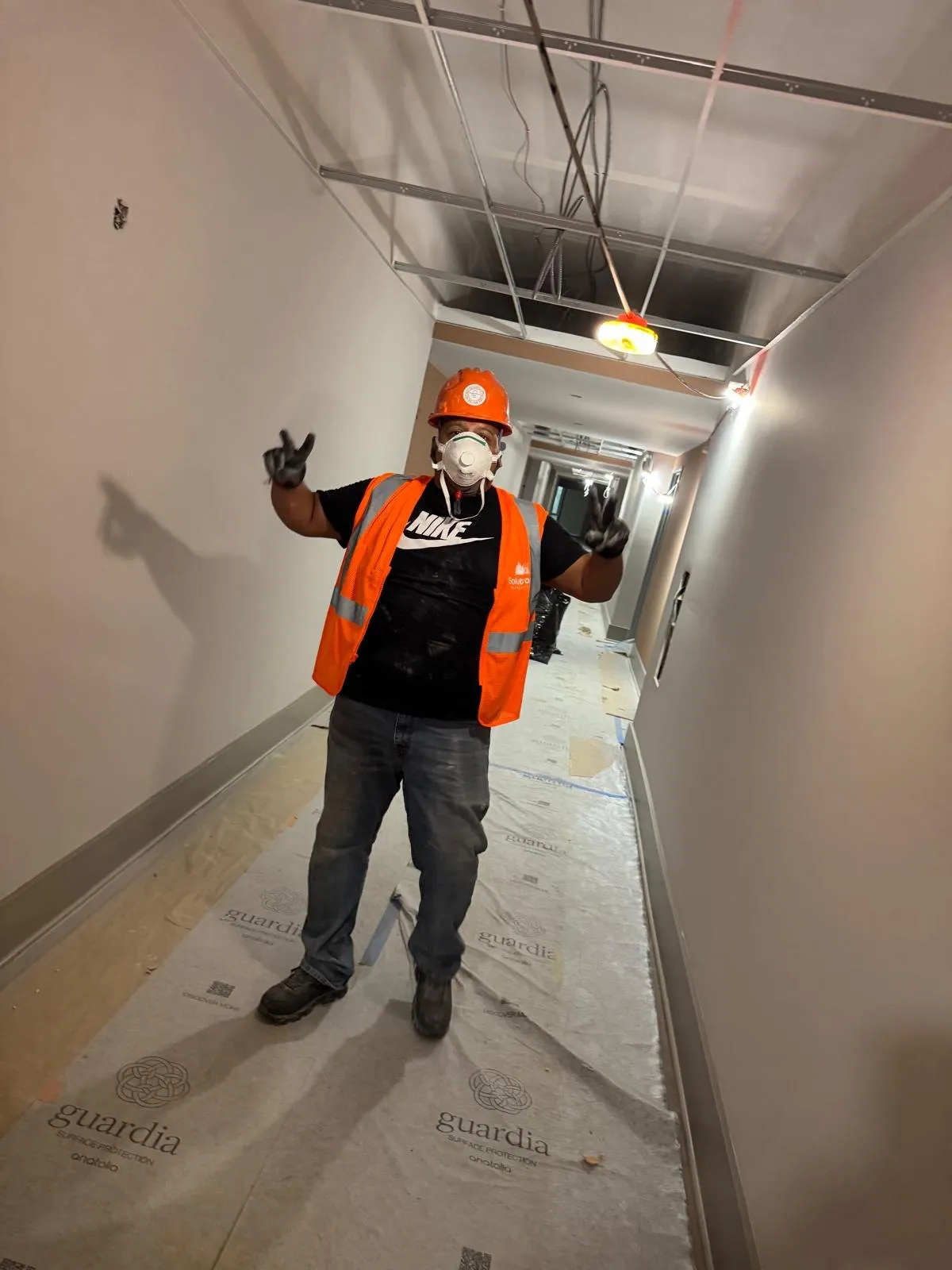 Solutions Cleaning crew member in a hard hat and N95 respirator giving peace signs in a corridor under construction