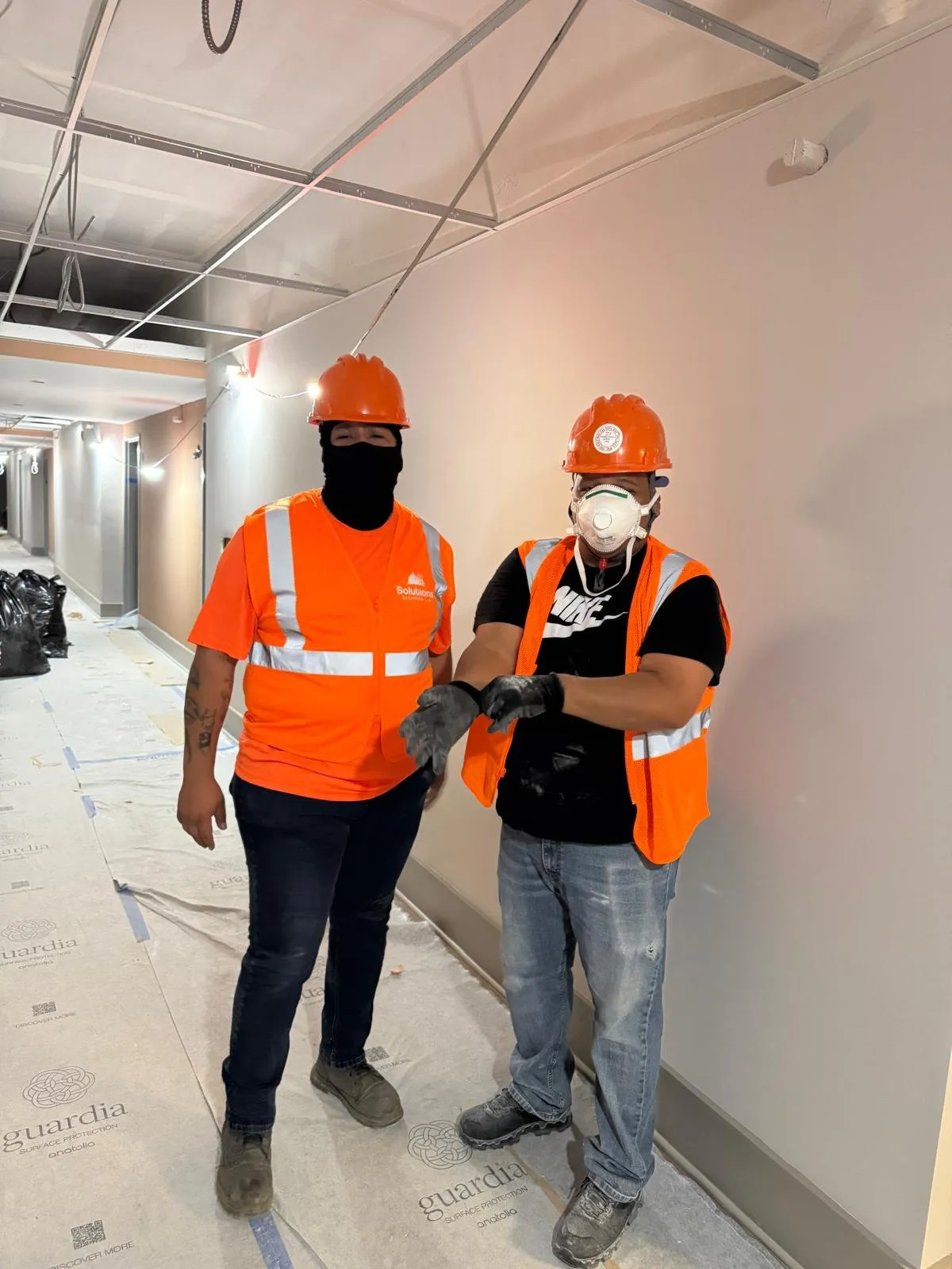 Two Solutions Cleaning crew members in hard hats, safety vests and respirators in a corridor with open ceilings and protected floors
