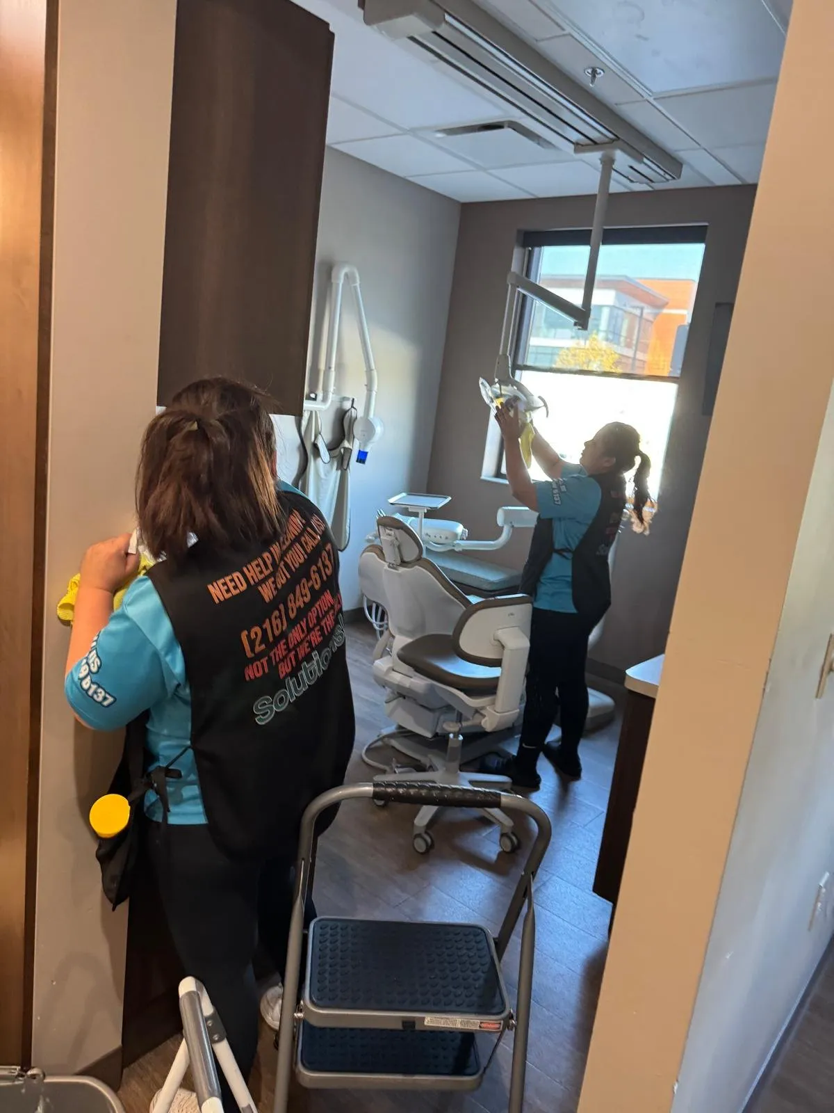 Two Solutions Cleaning team members cleaning a dental operatory, one dusting the overhead dental light