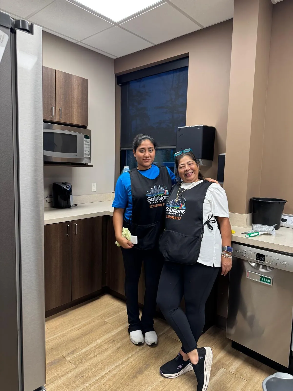 Two Solutions Cleaning team members in branded aprons smiling in a clean break room kitchen